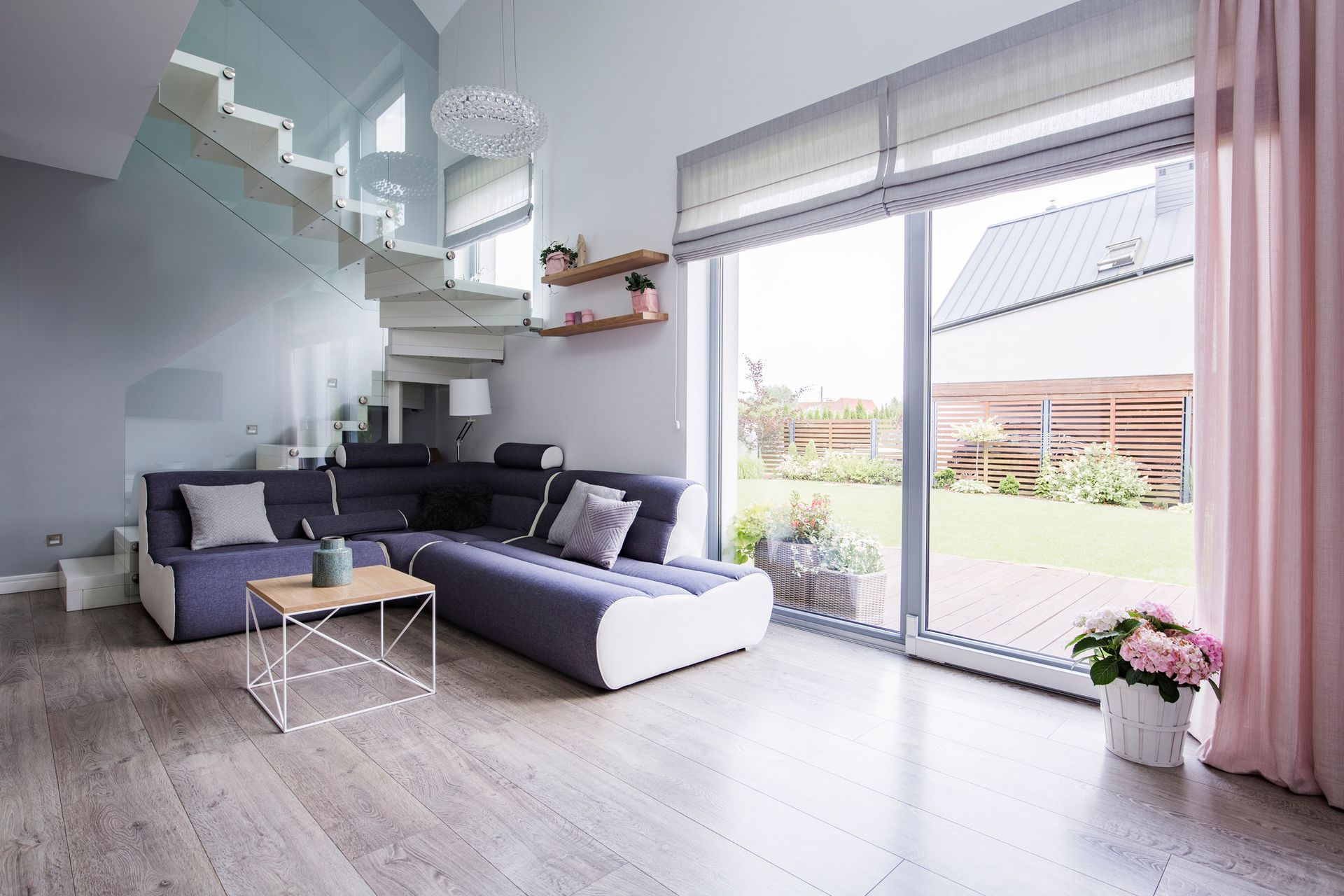 Modern living room with staircase, large windows, blue sofa, and pink curtains.