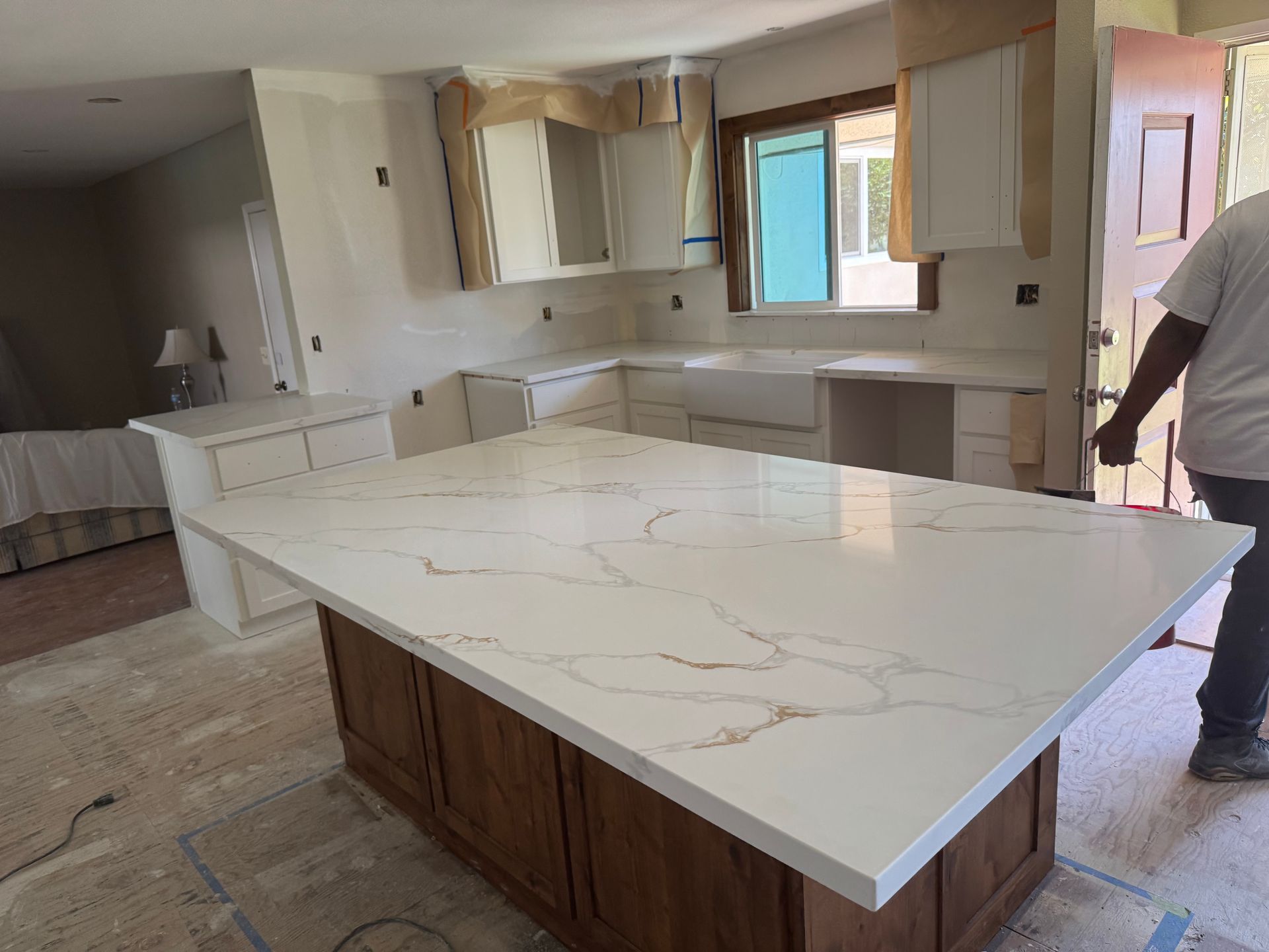 A kitchen under renovation featuring a large white quartz island with gold veining and a person standing to the right.