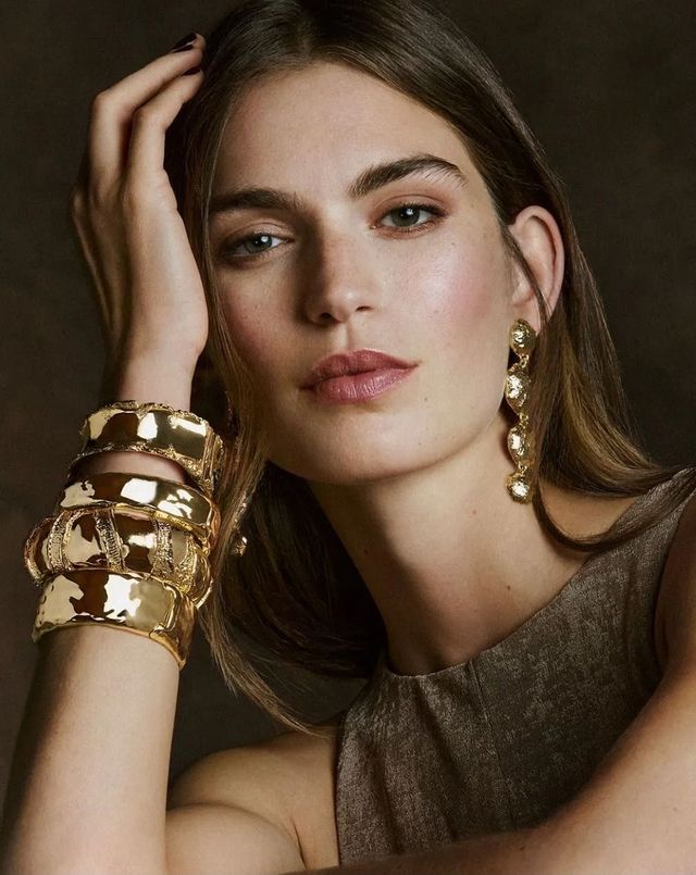 Ritratto di una donna che indossa gioielli d'oro, con la mano appoggiata sulla testa, su uno sfondo scuro.