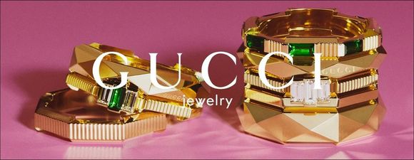 Espositore di gioielli Gucci con anelli d'oro e pezzi geometrici sovrapposti su sfondo rosa.