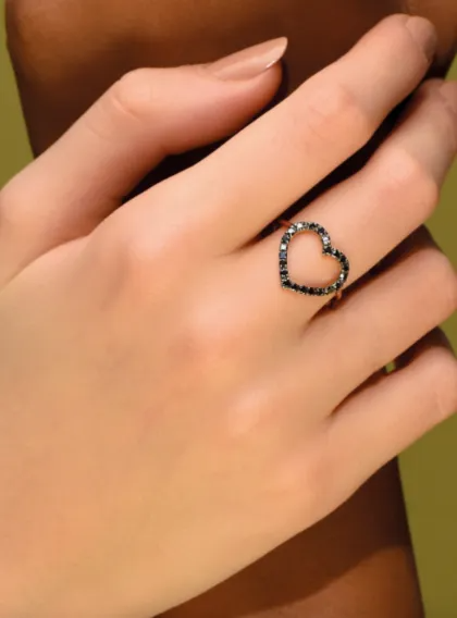 Mano con un anello nero a forma di cuore su uno sfondo beige tenue.