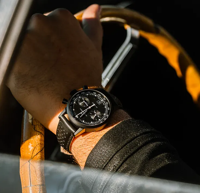 Primo piano di un polso con un orologio nero mentre si tiene il volante di un'auto sotto una calda luce solare.