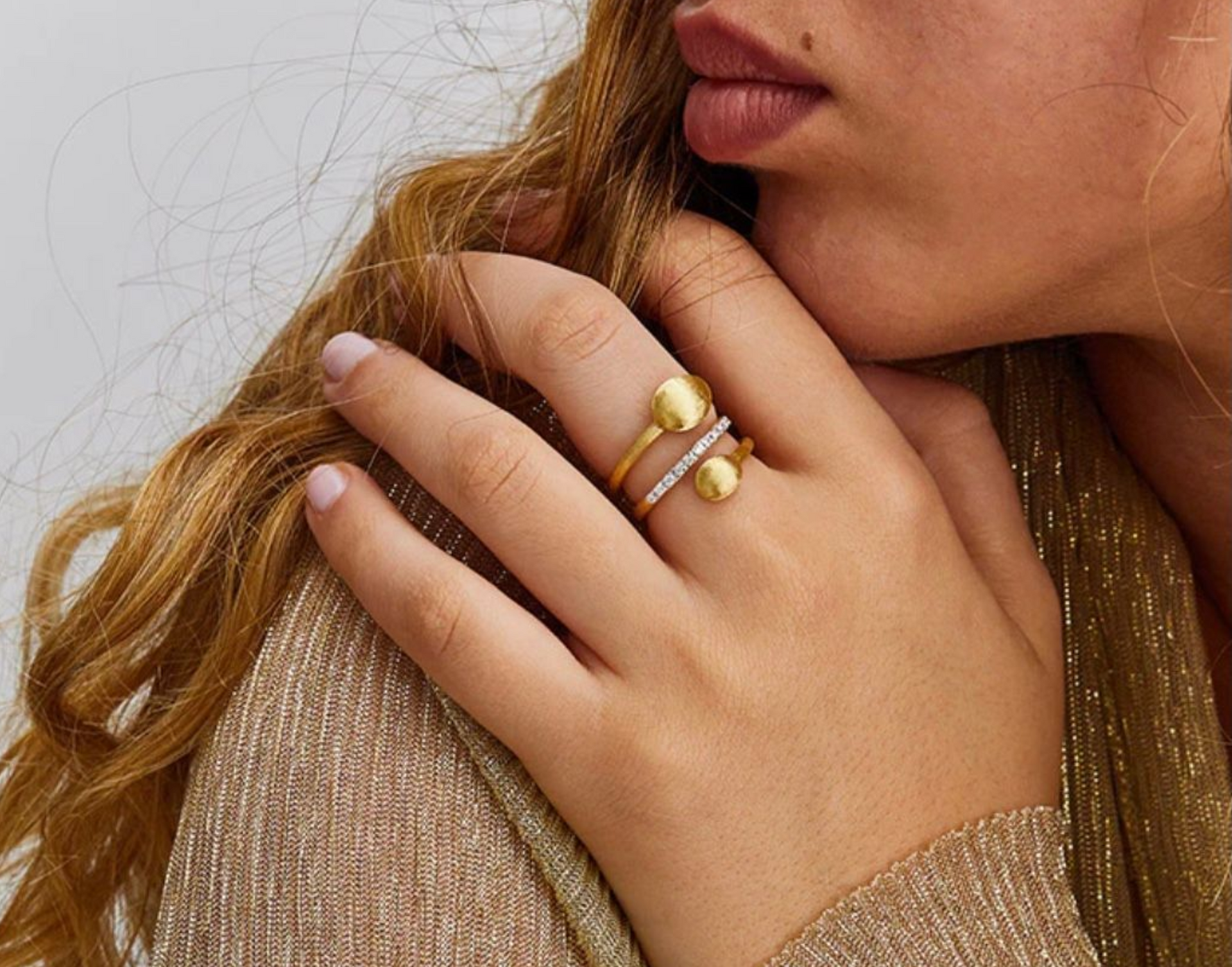 Primo piano di una mano con anelli d'oro e di perle appoggiata su capelli mossi e un maglione beige.