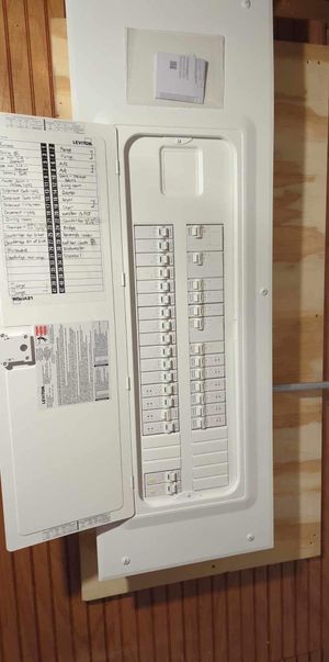 White electrical panel or breaker box mounted on a wood wall next to a small paper notice.