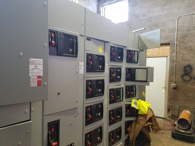 Electrical control panel room with gray switchgear cabinets and a worker in a yellow shirt crouching beside them