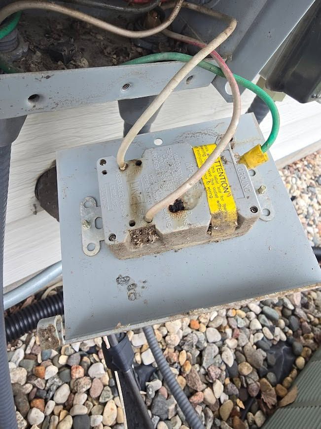 Rusty electrical junction box with exposed wires mounted on a gray metal plate outdoors.