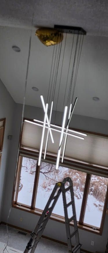 Modern LED chandelier hanging from the ceiling near a window; a ladder stands below during installation.