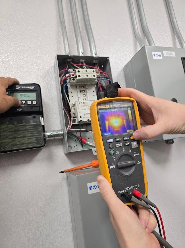 Technician using a thermal camera to inspect an open electrical panel with exposed wiring and breaker boxes.