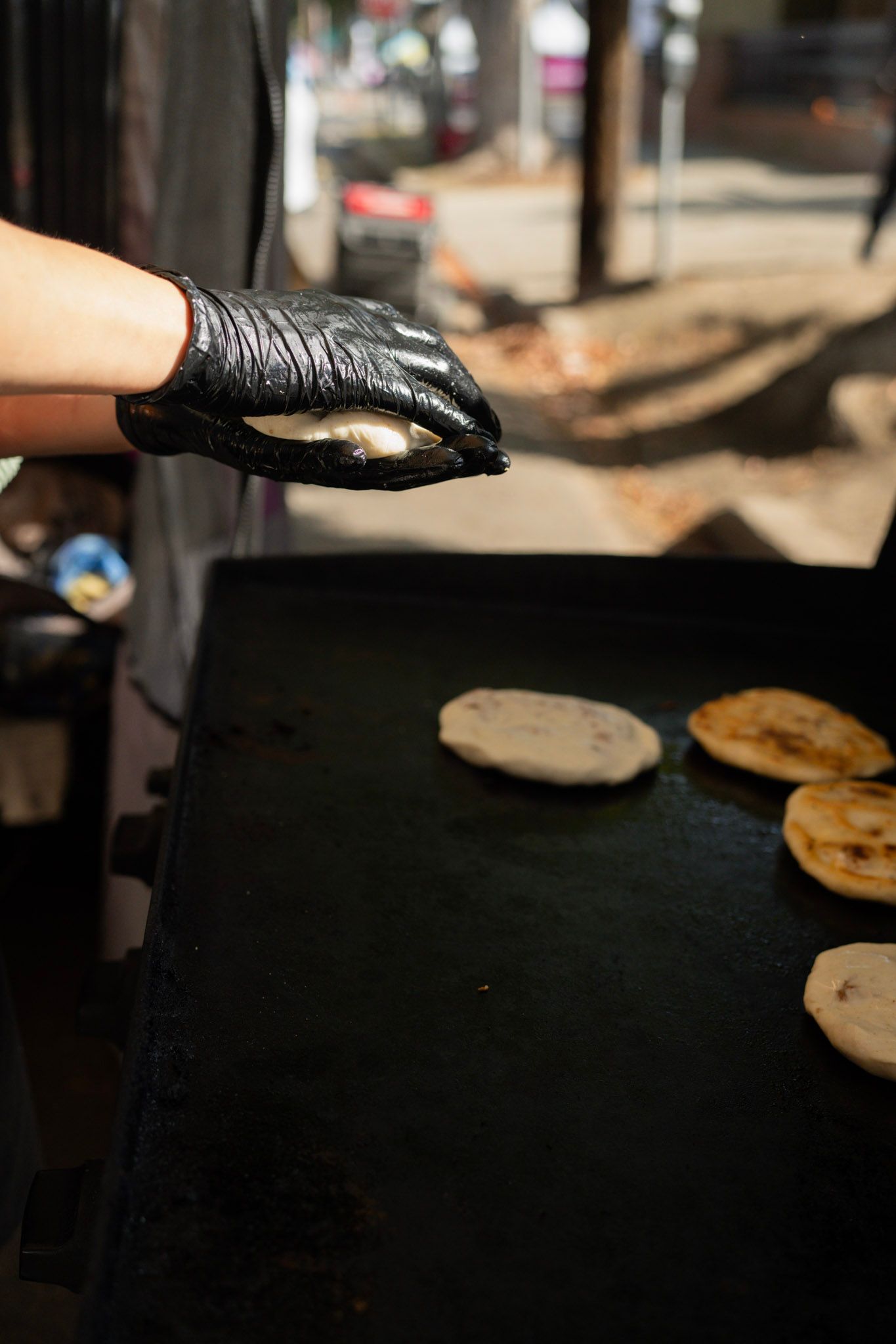Person in black gloves placing a filled flatbread on a hot griddle with other cooking flatbreads.