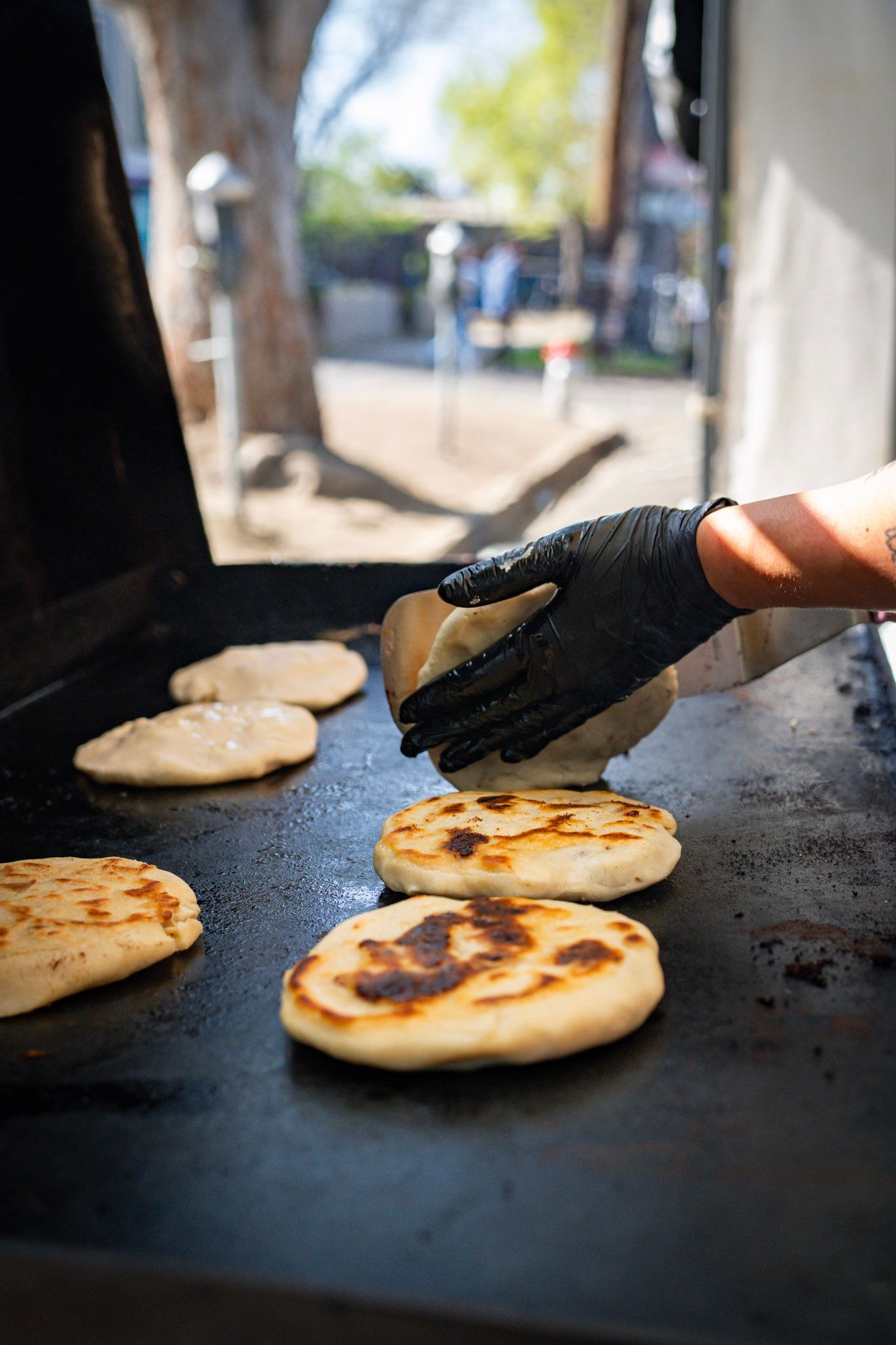 Person in black gloves presses down on cooking arepas on a hot griddle at an outdoor food stall.