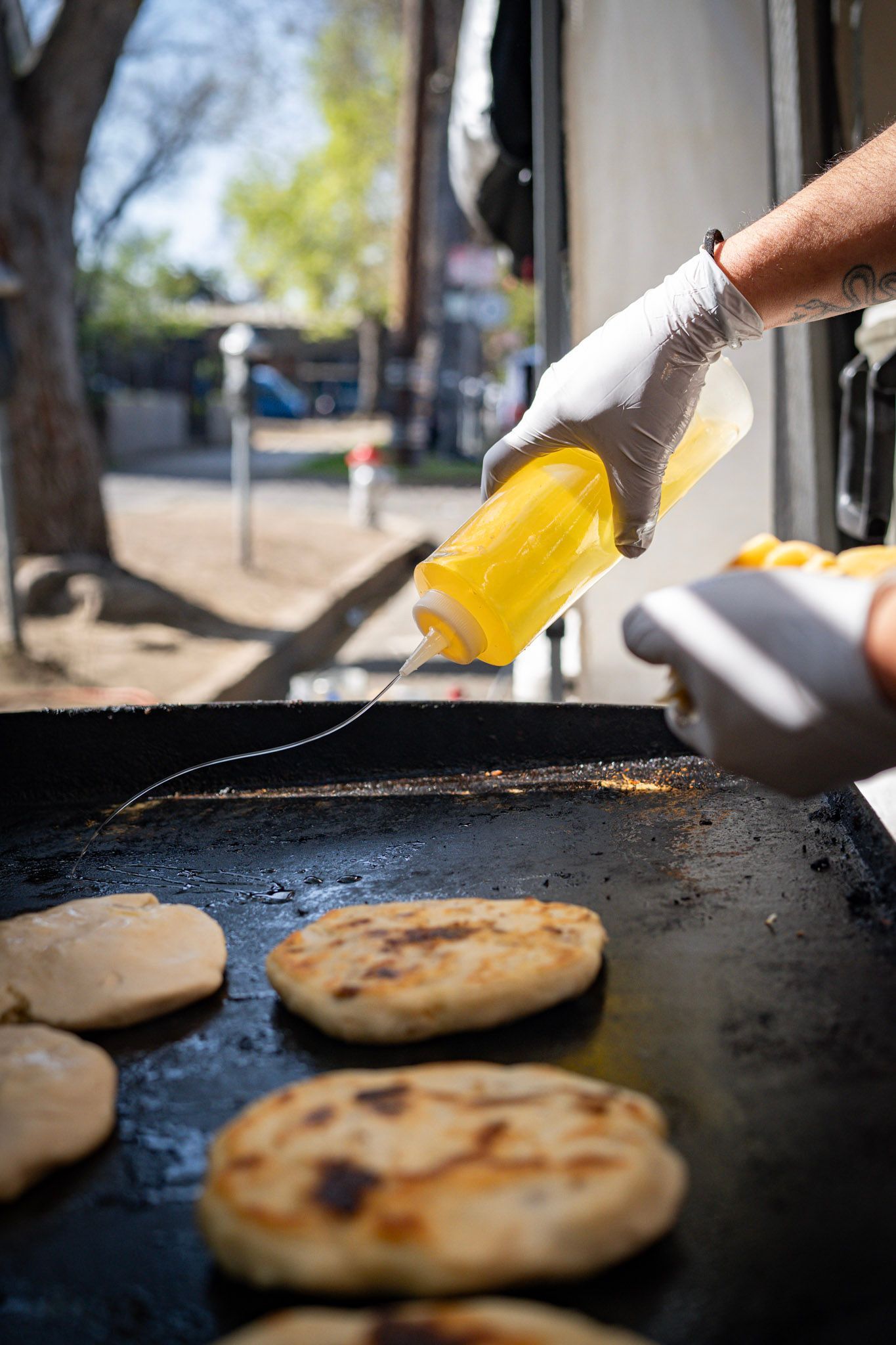 A person in gloves oils flatbreads on a hot griddle at an outdoor food stand.