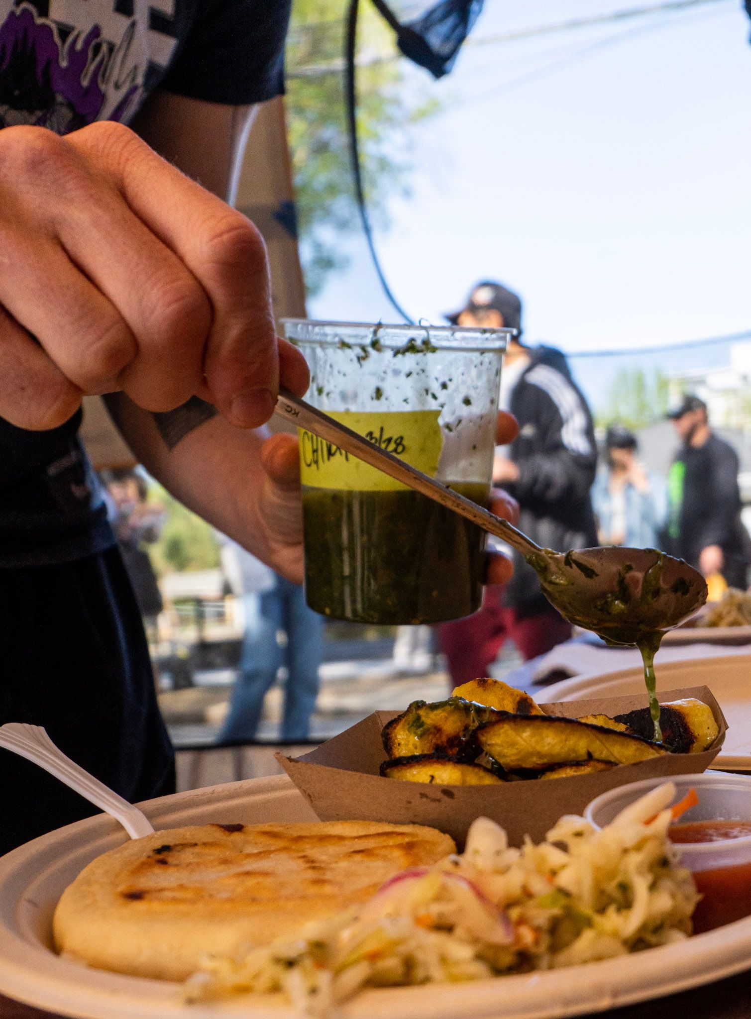 Person pouring green sauce over food: grilled squash, pupusa, and slaw, outdoors.
