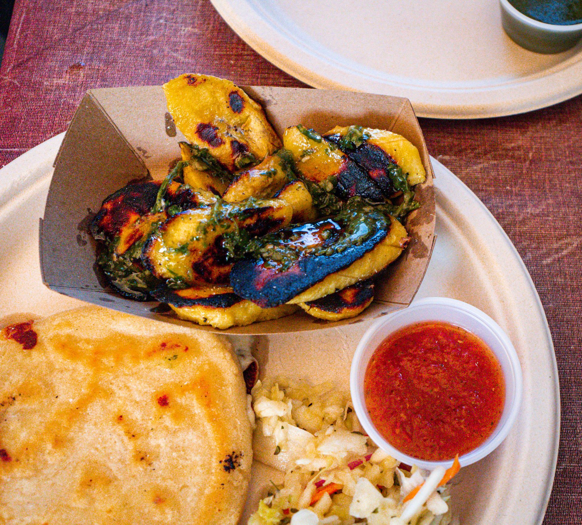 Plate of fried plantains with green sauce, slaw, salsa, and a pupusa on a wooden table.