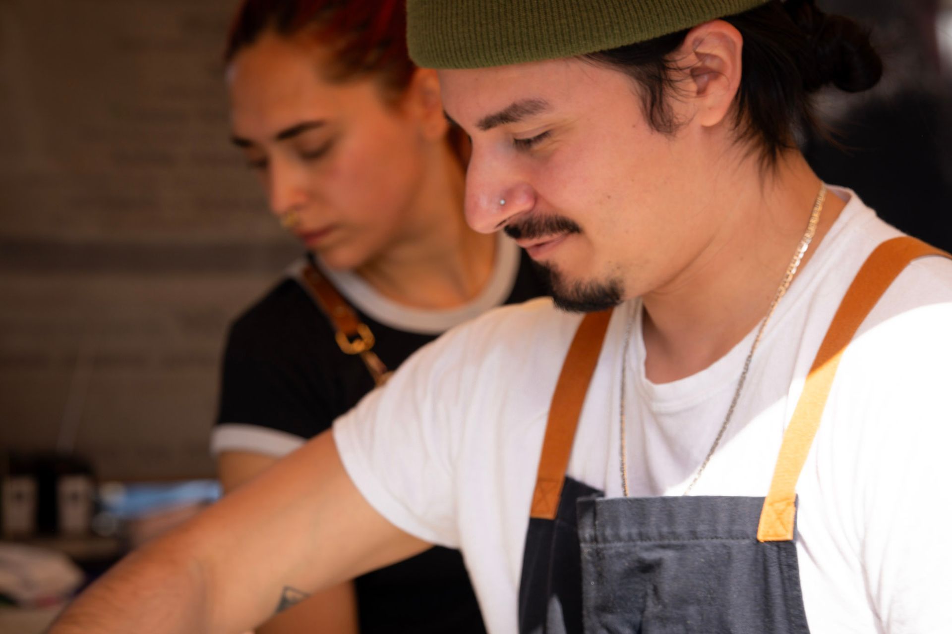 Two people, a man and a woman, work at a food stand. Man in white shirt, black apron, green beanie. Woman in black shirt.