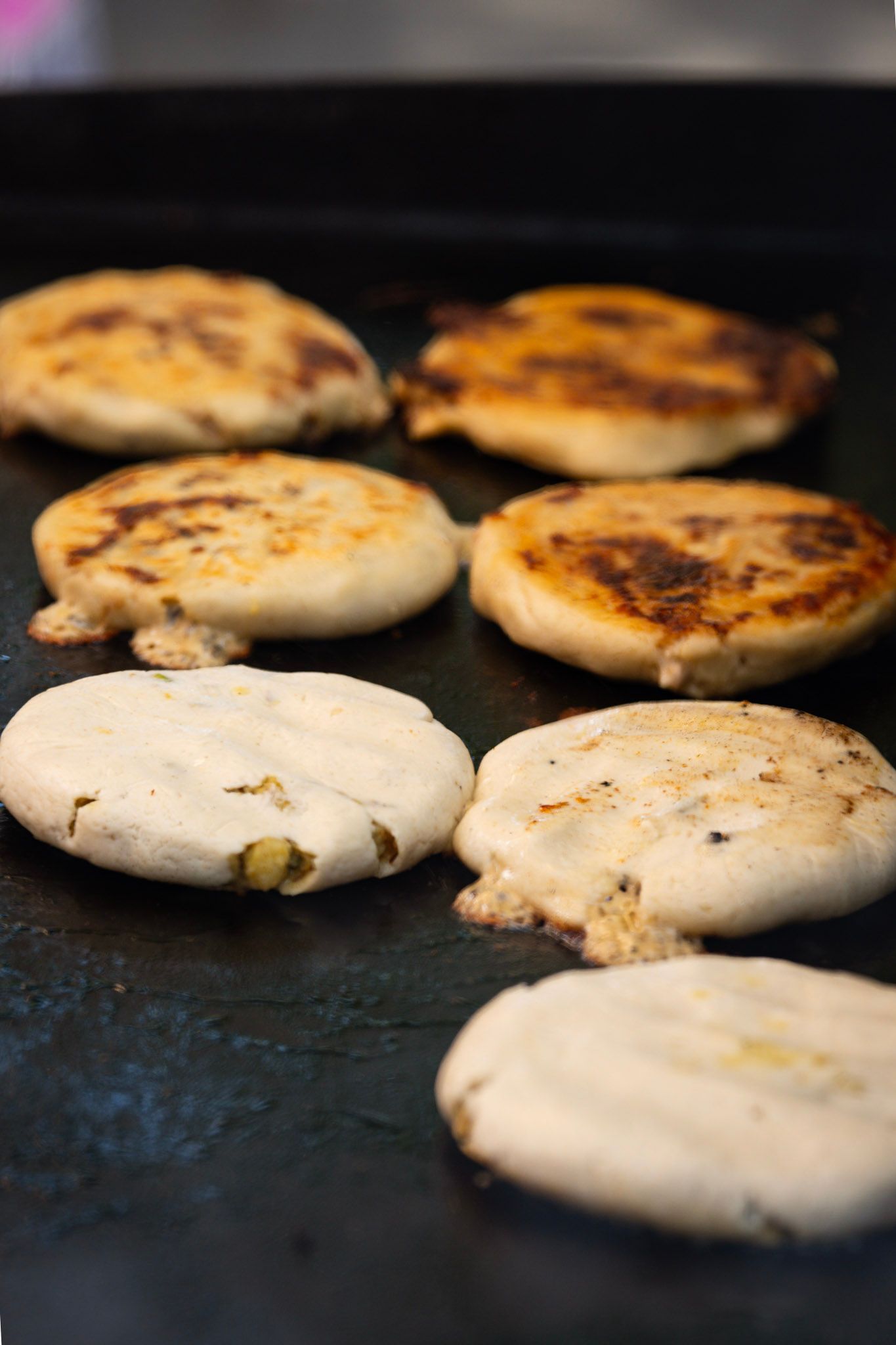 Arepas cooking on a black griddle, some with melting cheese, golden-browned.