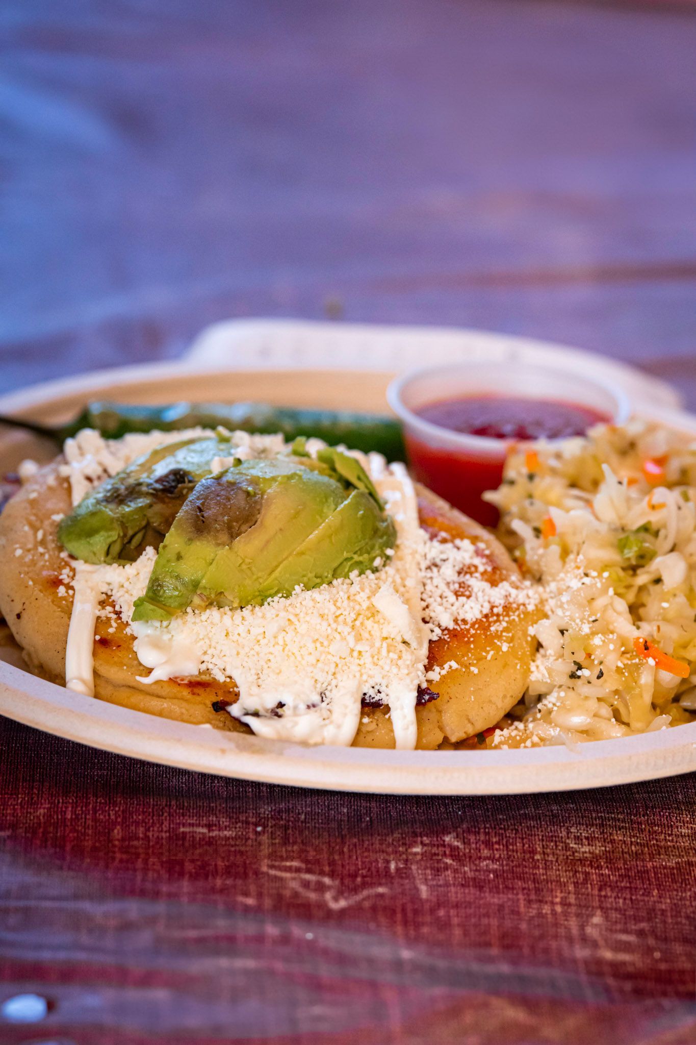 Plate with pupusa topped with avocado, cheese, and salsa, with curtido and hot sauce.