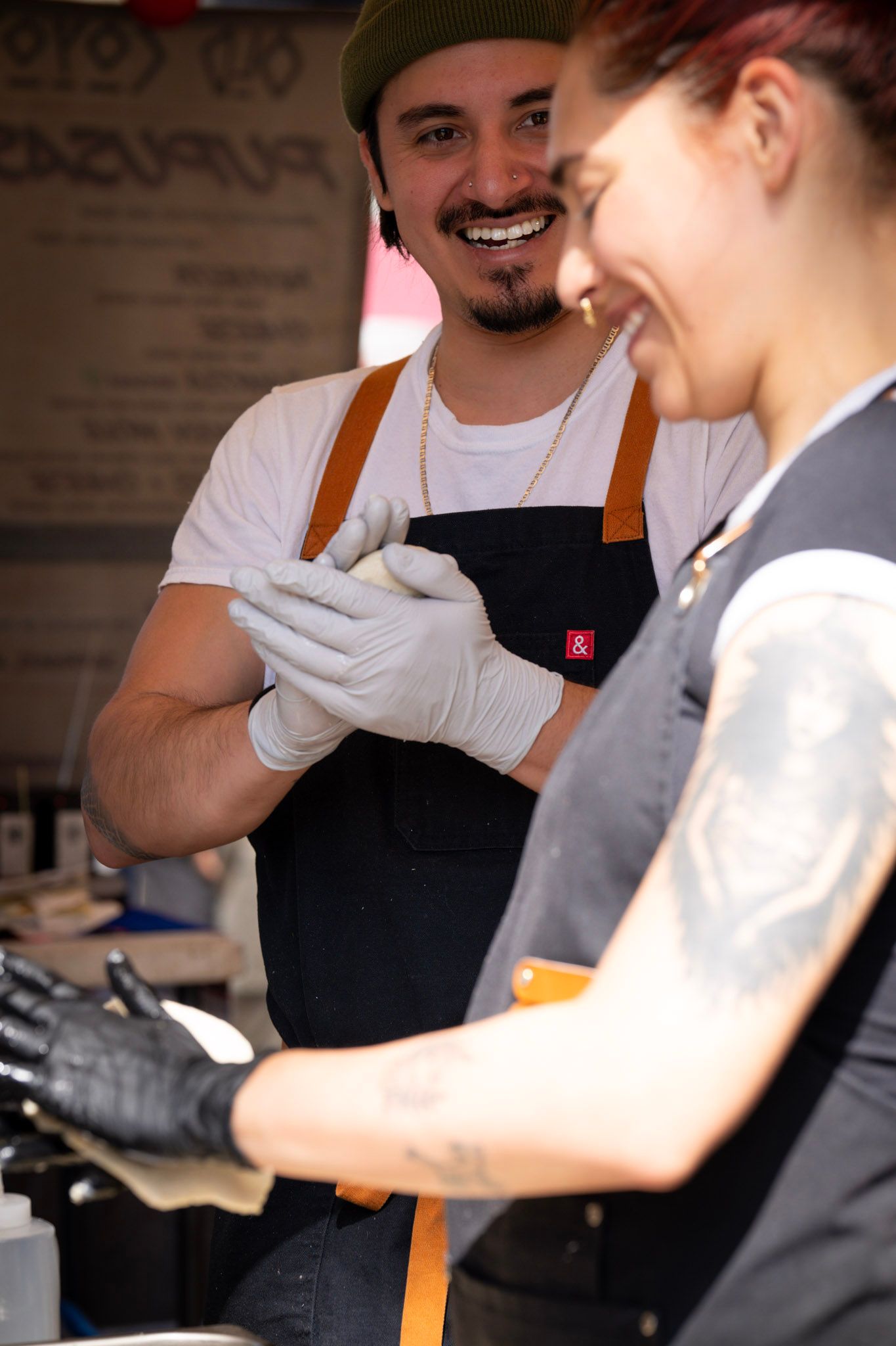 Two chefs smiling, wearing aprons and gloves, working in a food stall.