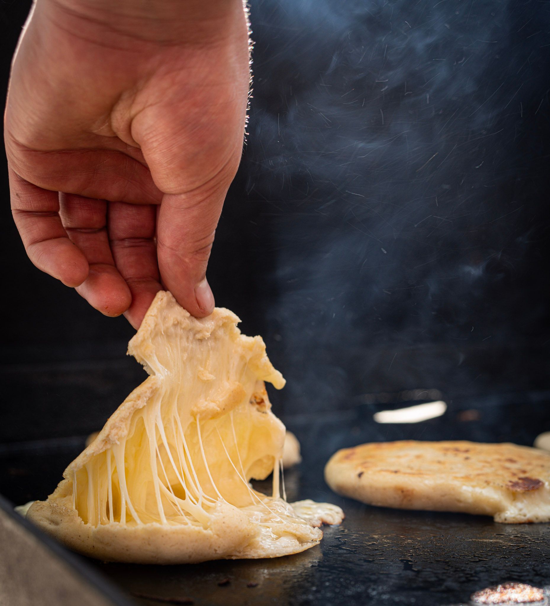 Hand lifting grilled arepa, cheese melted, on a black griddle.