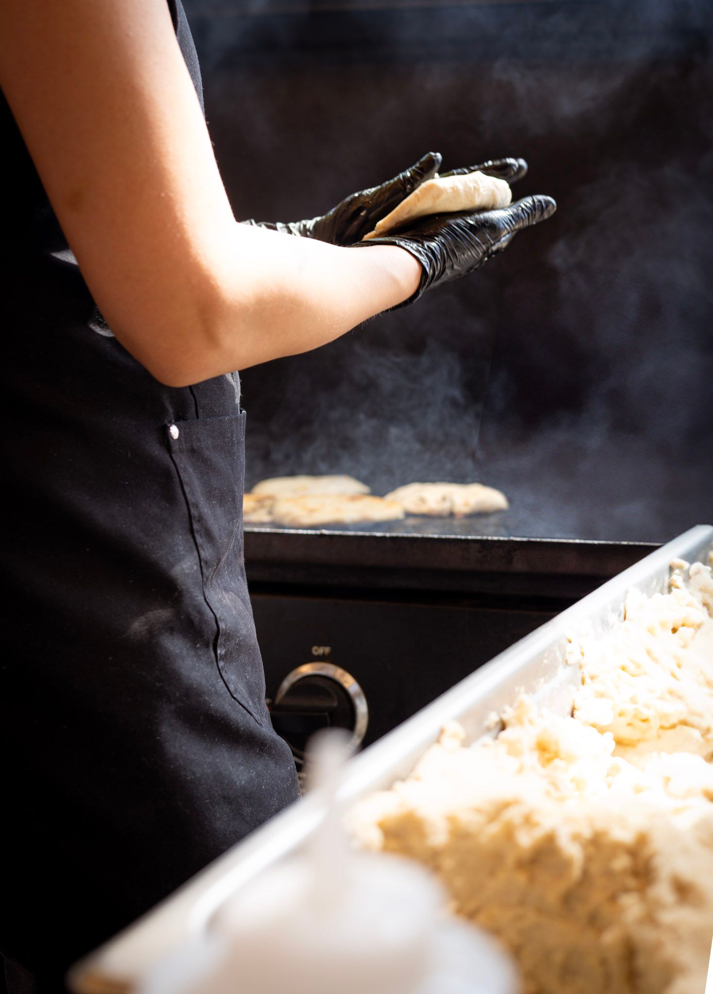 Person in black apron and gloves cooking patties on a grill, with a tray of food in the foreground.
