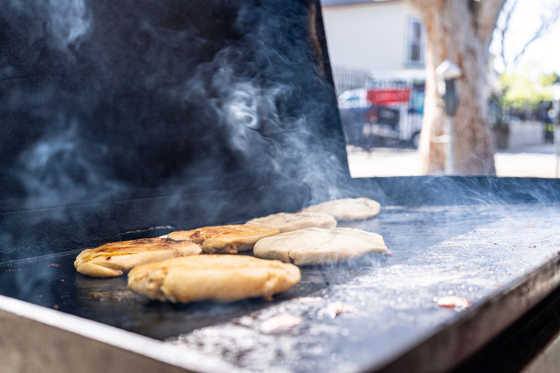 Patties sizzling on a hot griddle, smoke rising. Outdoors, near a street.