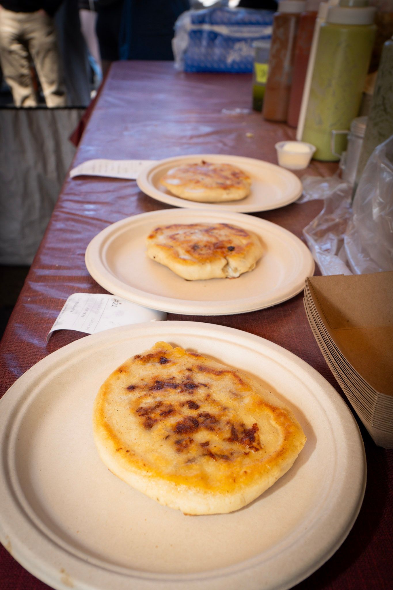 Three golden-brown arepas on plates sit on a red table at a food stall.