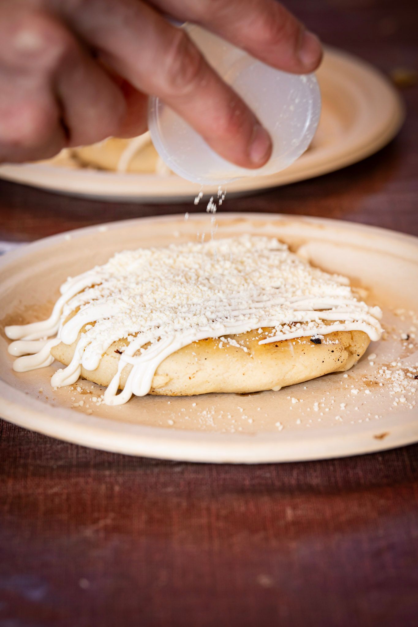 Person sprinkles cheese over an arepa topped with white cheese on a plate.