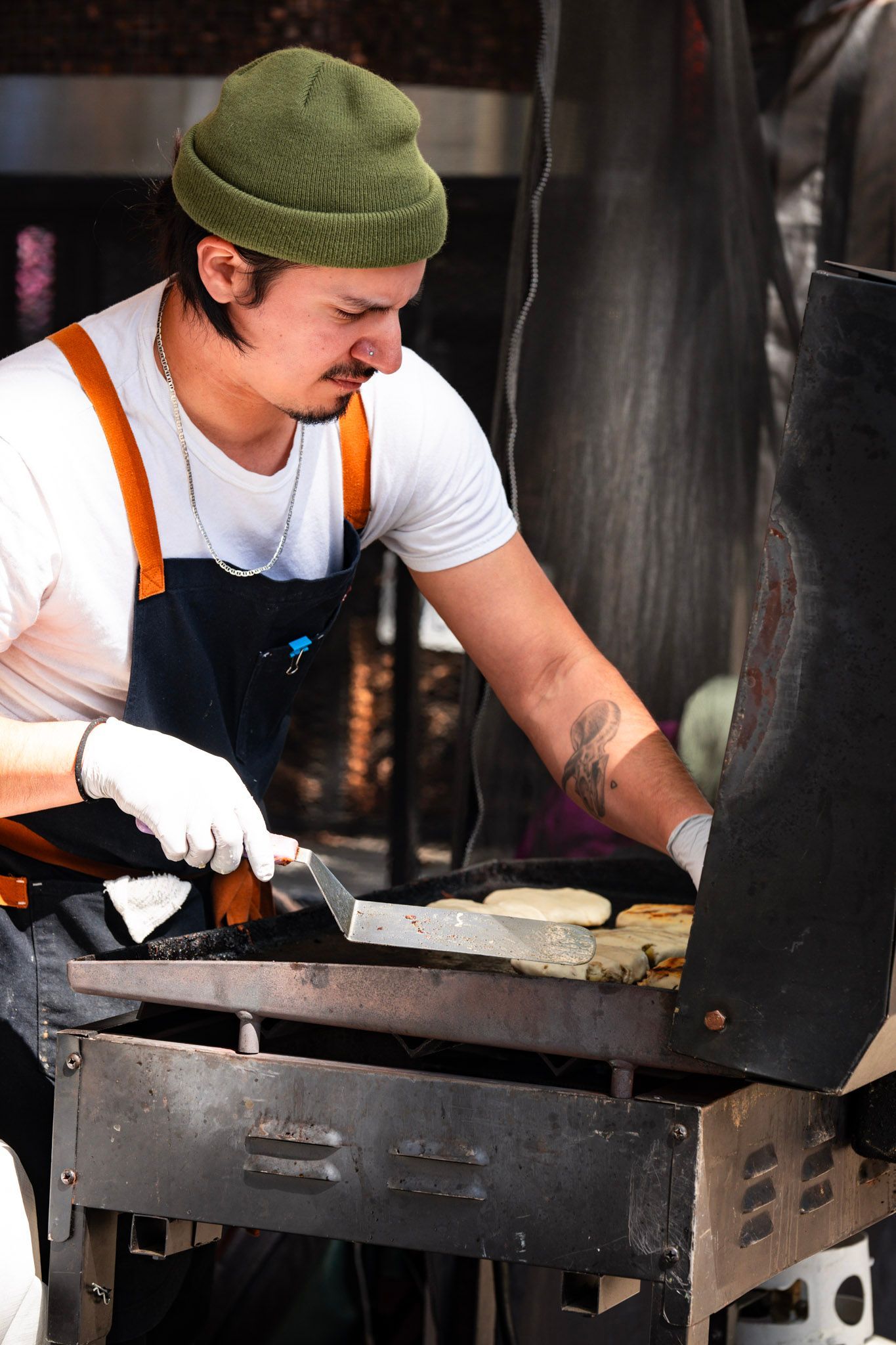 Man grilling food outdoors, wearing a green beanie, apron, and gloves.