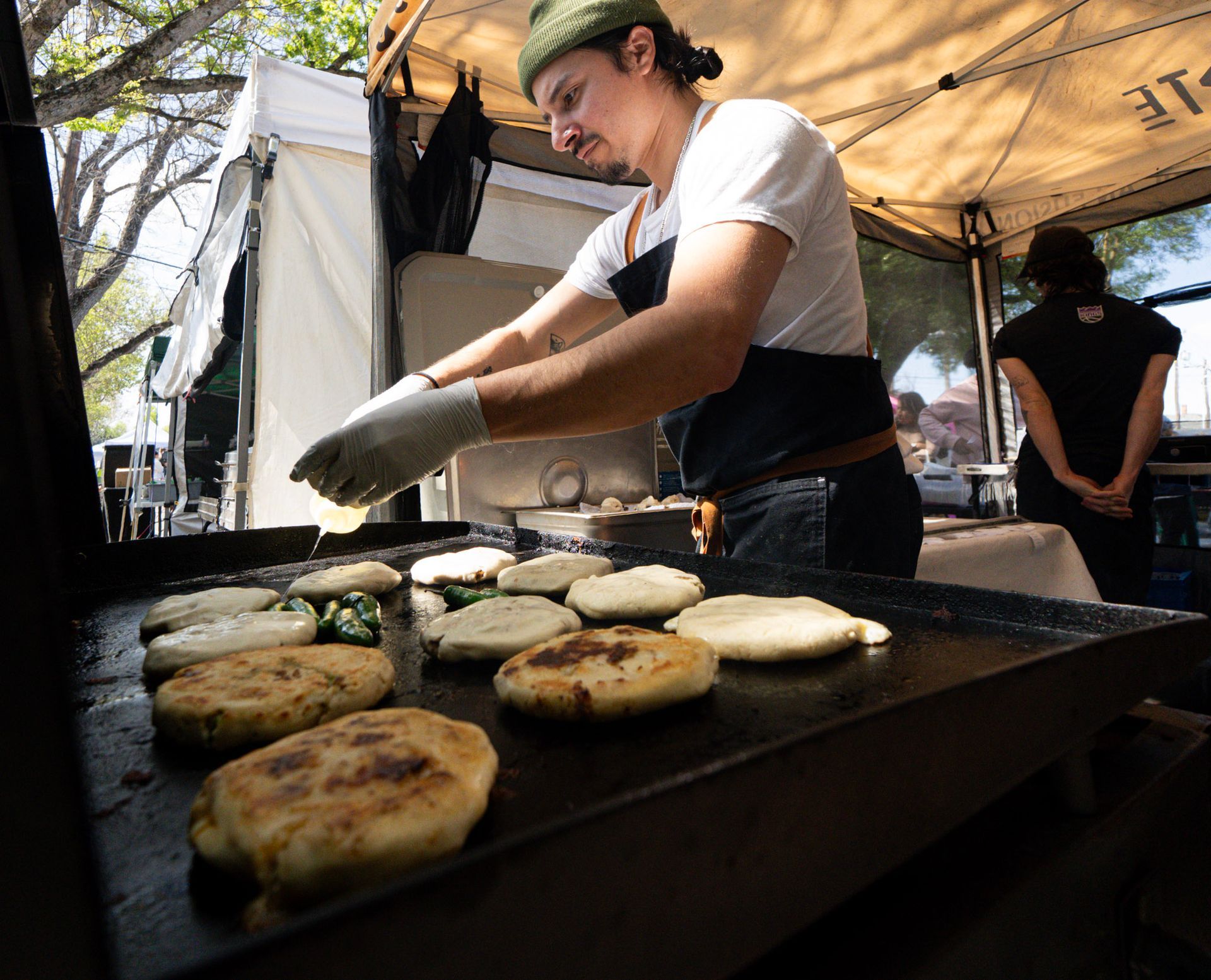 Chef grilling arepas at an outdoor food stall. Man in apron and gloves flips golden-brown patties on a large griddle.