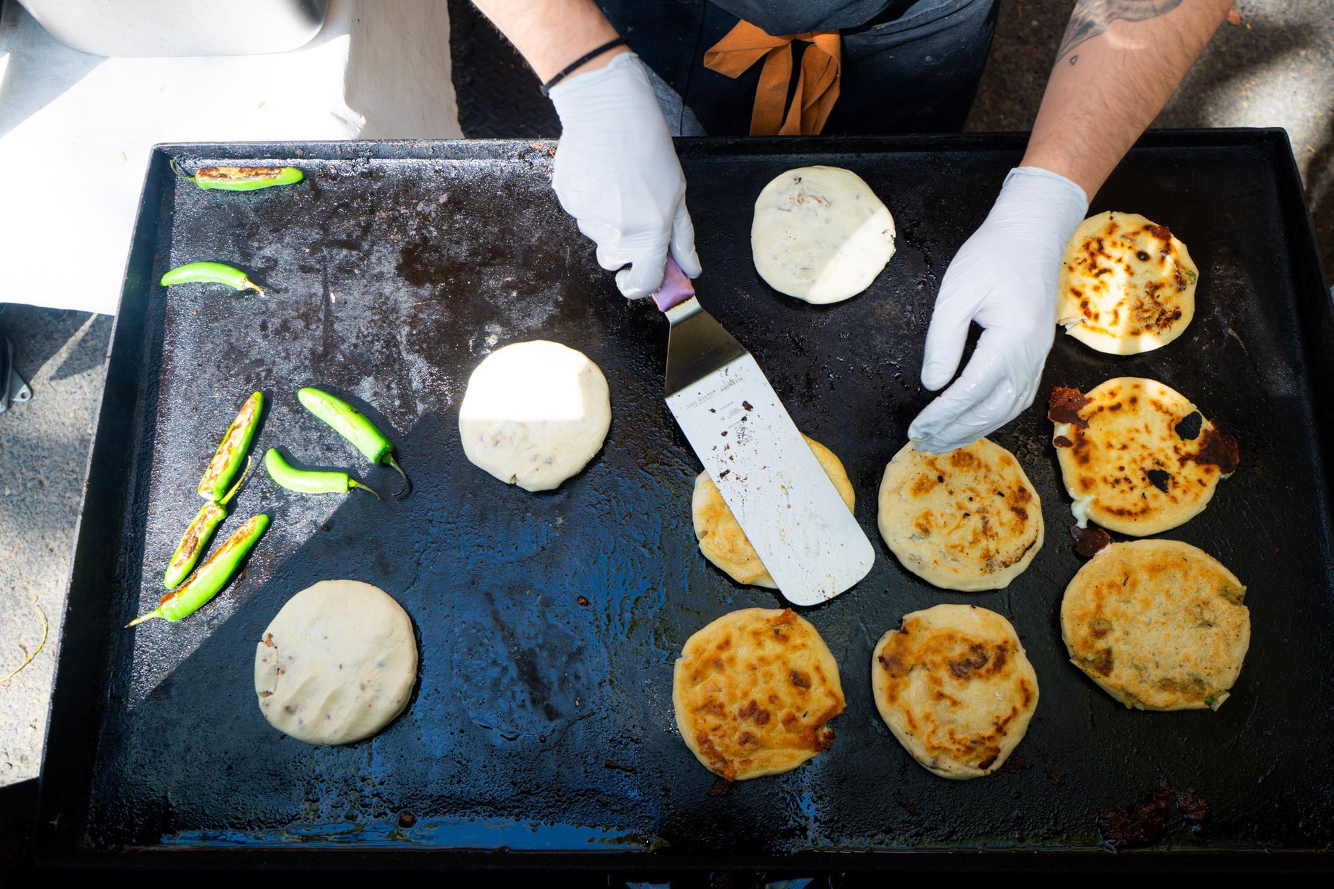 Person grilling arepas and peppers on a flat-top griddle; close-up of hands in gloves flipping food.