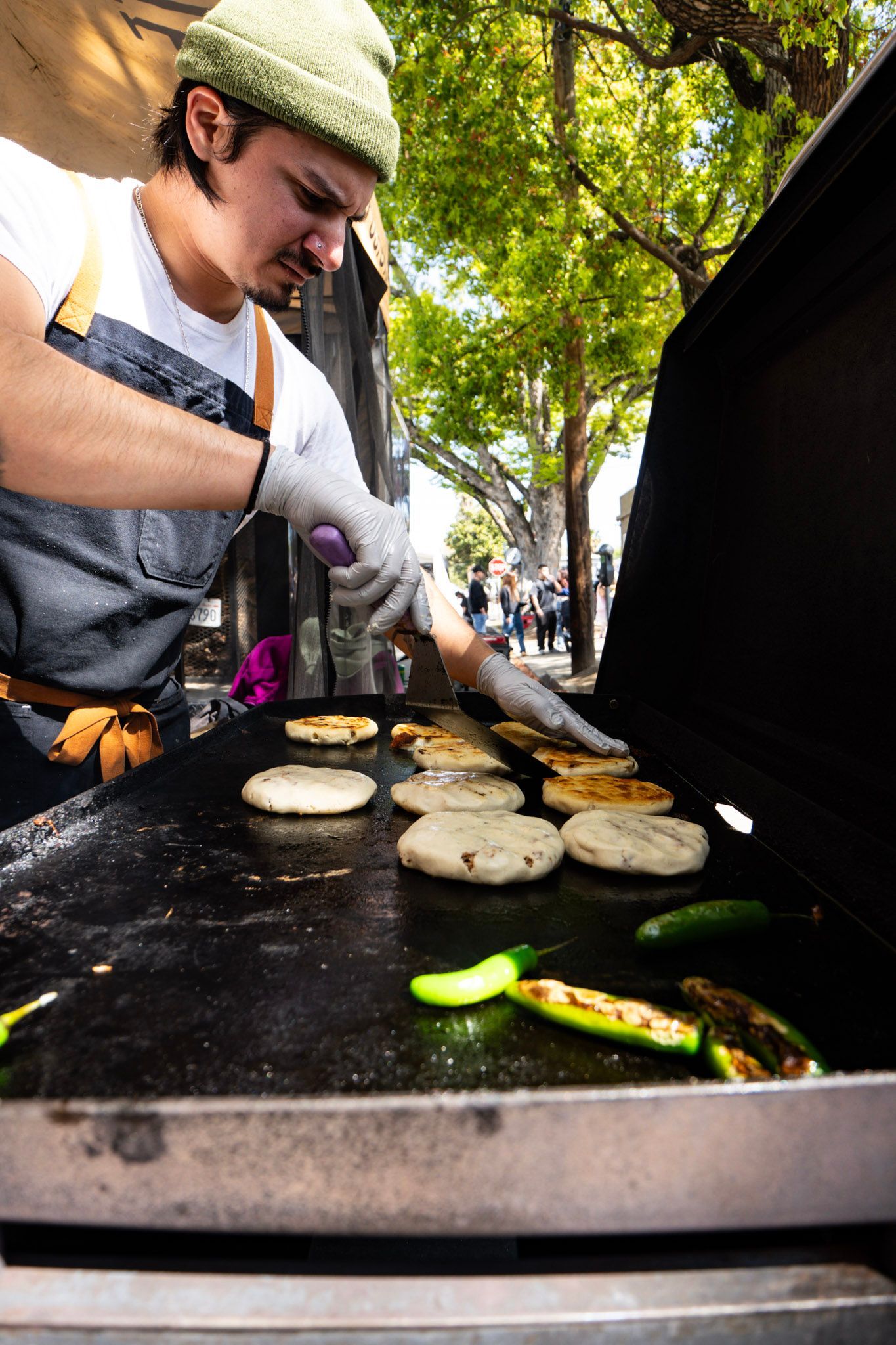 Man grilling arepas and jalapeños on a flat top grill outdoors. He wears a beanie, apron, and gloves.