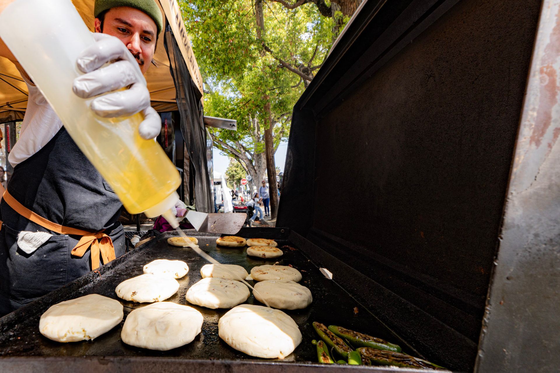 Man pouring oil onto arepas on a griddle at an outdoor food stall.