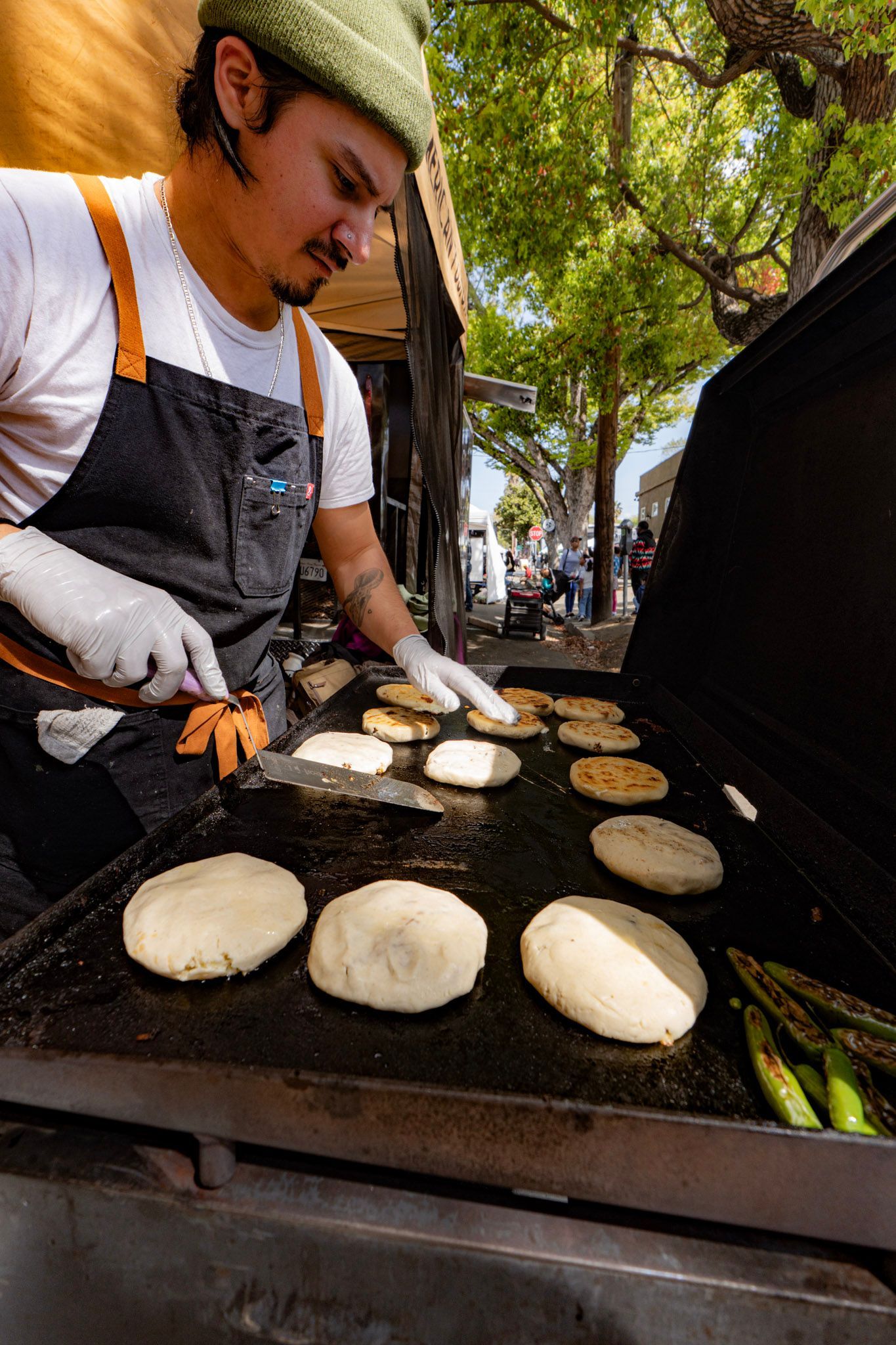Chef grilling tortillas on a flat top. Outdoors, wearing apron, gloves, and a green hat.