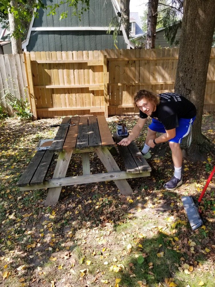 A man is painting a wooden picnic table in a backyard.