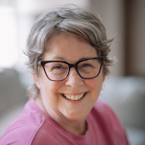 A woman wearing glasses and a pink shirt is smiling for the camera.