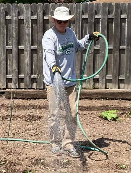 A man wearing a green bay packers shirt is watering a garden with a hose