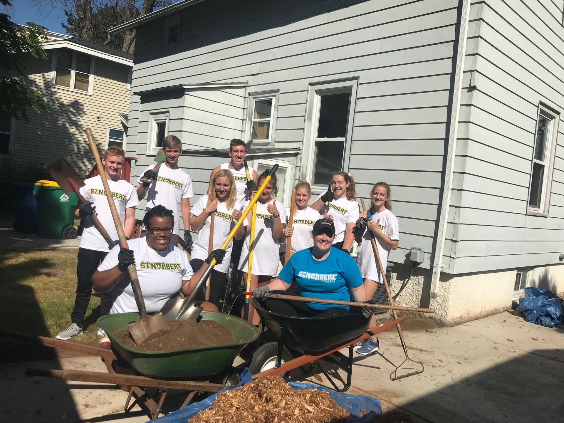 A group of people are standing in front of a house holding shovels and a wheelbarrow.
