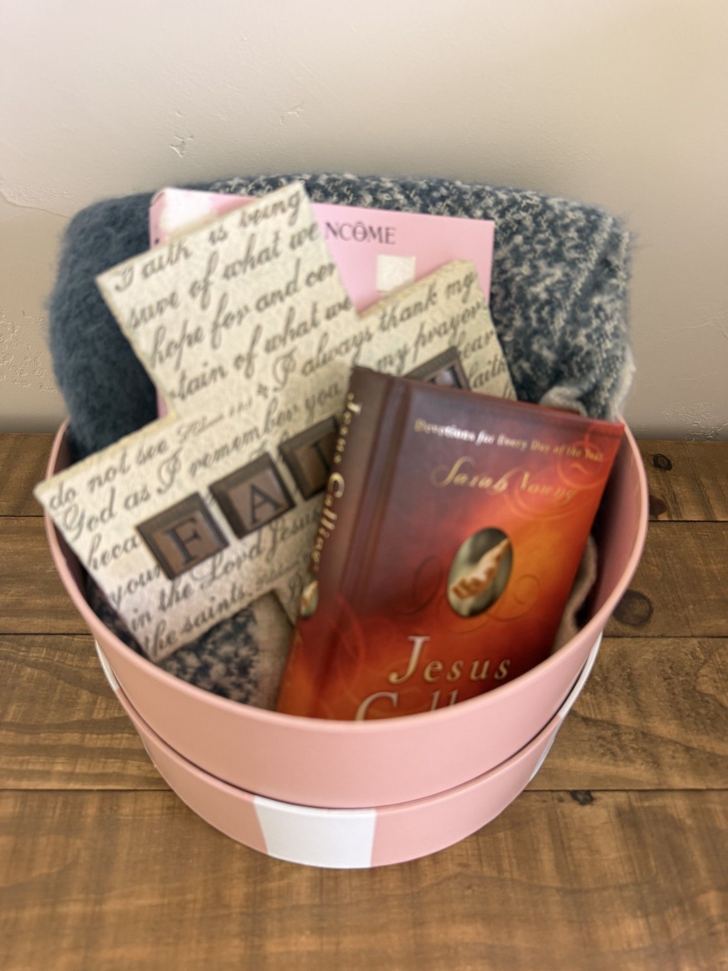 Pink bucket gift basket with candles, a sign, and a Jesus-themed book on a wooden table