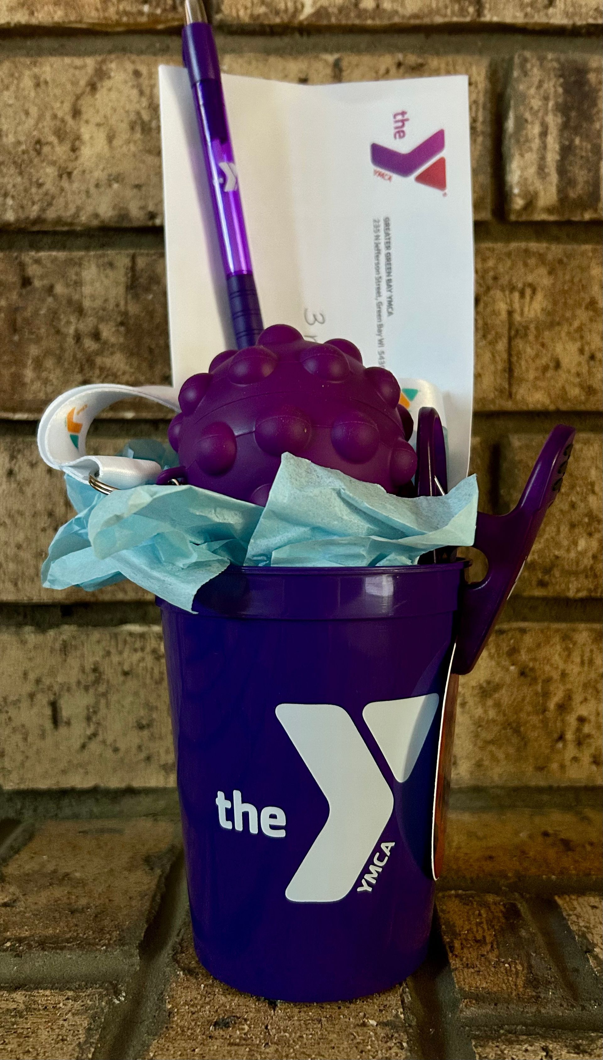 Purple cup with a white “Y” logo holding a purple pen, blue tissue paper, and a purple grape-shaped item on a stone wall.