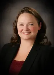 Woman with brown hair smiles, wearing a black blazer and red top. Dark background.