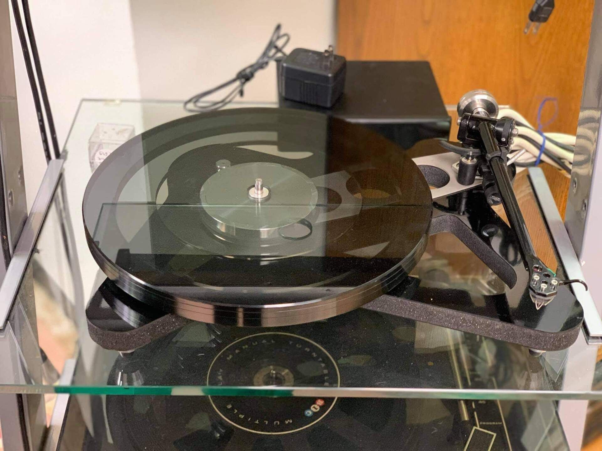 Glass turntable with vinyl record, tonearm, and a small black power adapter on top of a cabinet.