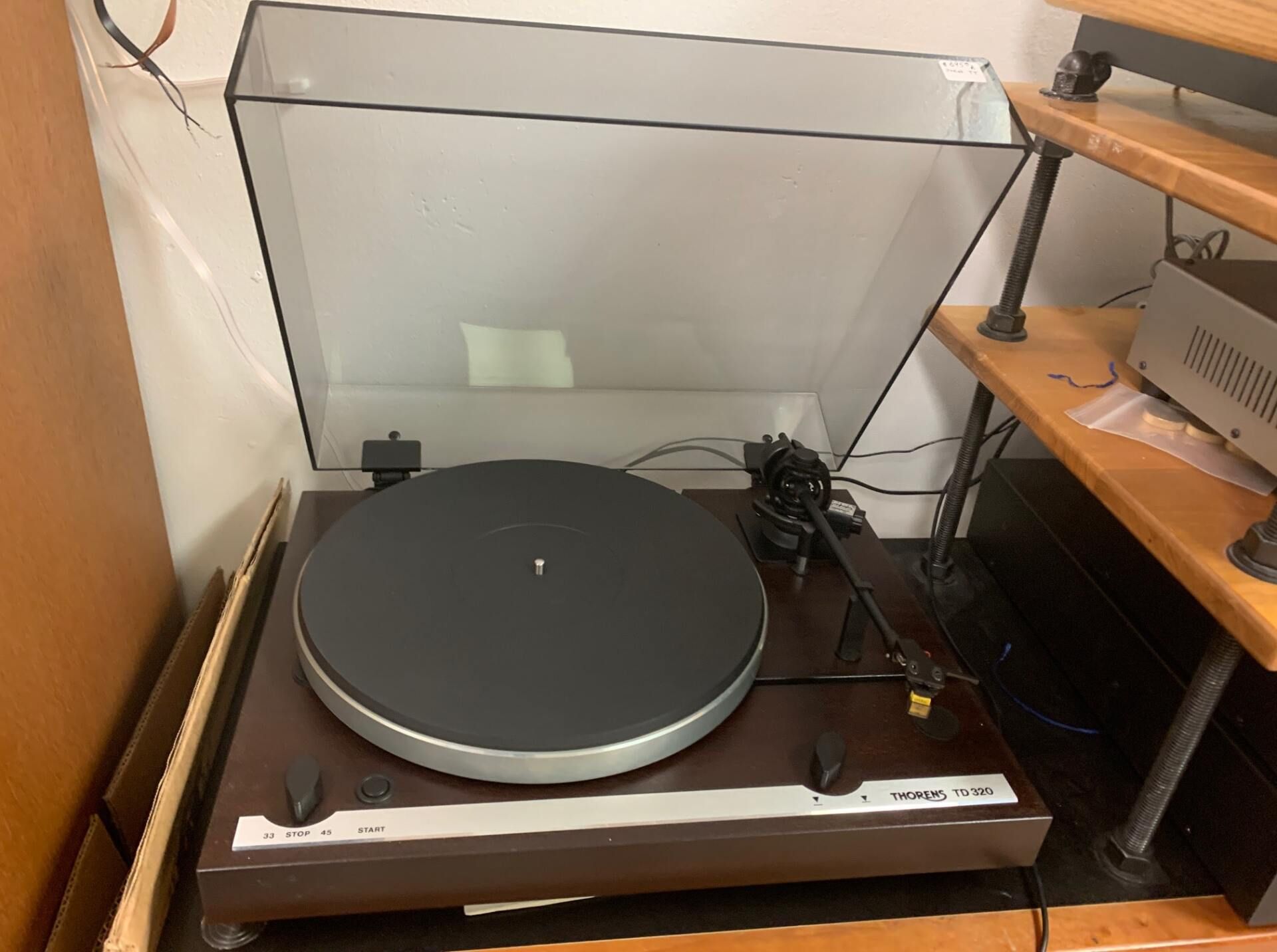 Turntable on a wooden shelf beside a computer monitor and speakers, with a clear dust cover open.