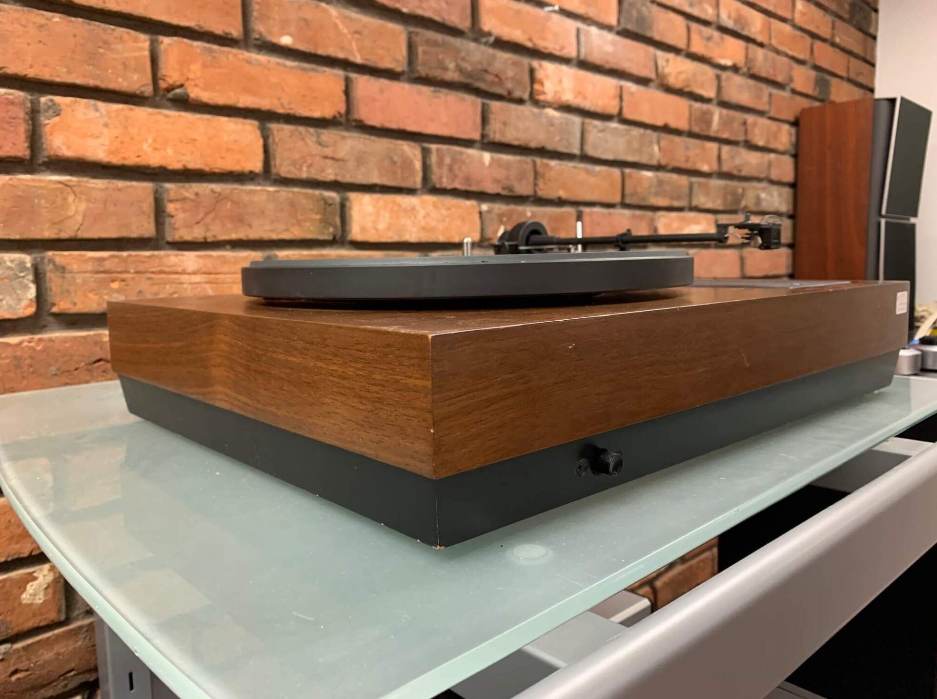 Wooden turntable with black platter on a glass shelf, set against a brick wall