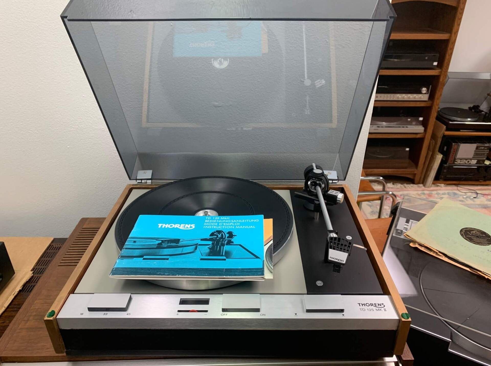 Turntable with dust cover open, a blue manual on the platter, and a tonearm on a wooden table