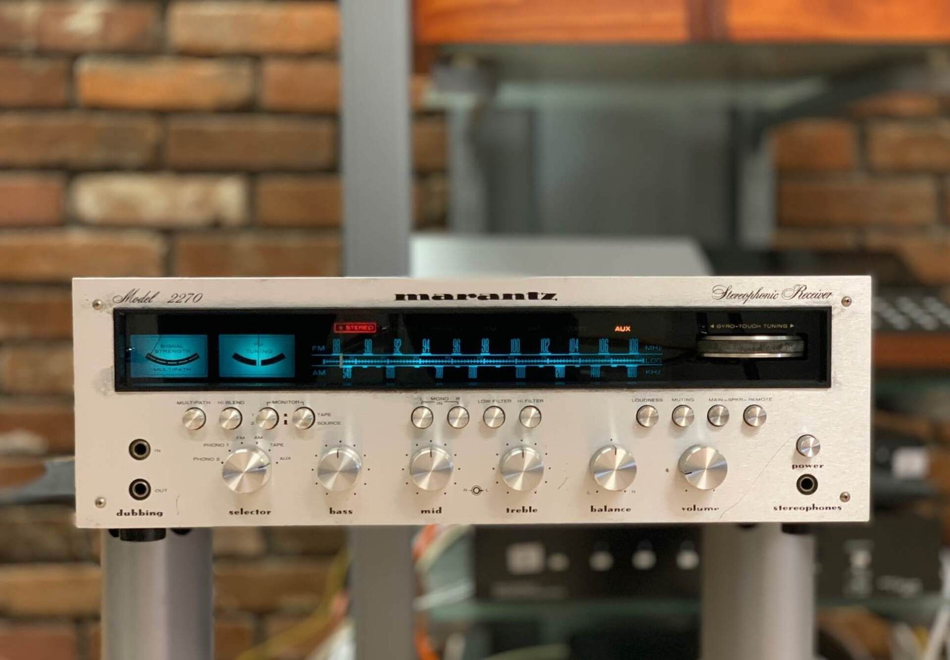 Vintage silver Marantz stereo receiver with blue-lit display on a shelf, against a brick wall.