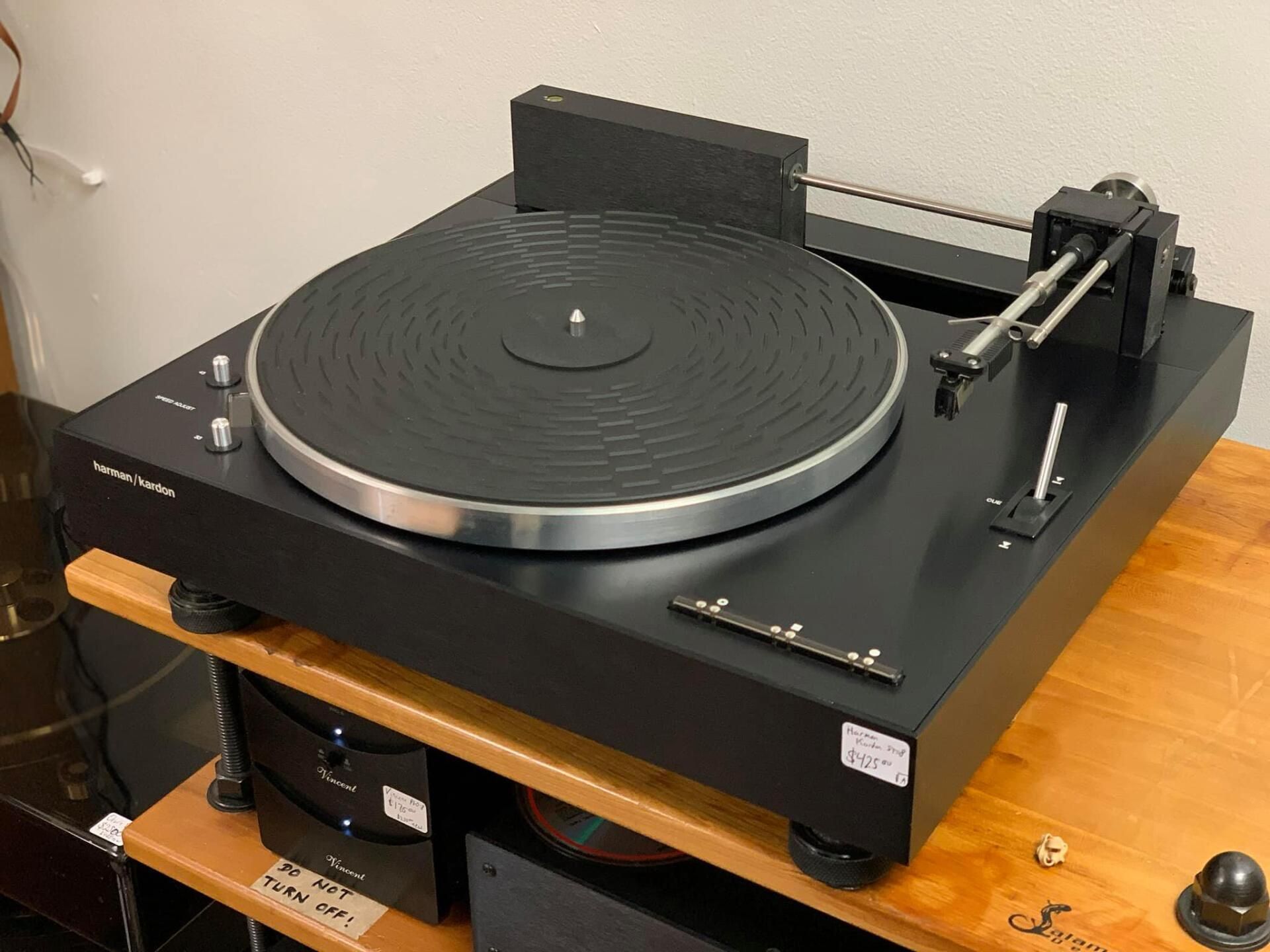 Black vinyl turntable on a wooden shelf with a tonearm and dust cover raised.