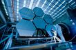 Large hexagonal mirrors, part of a telescope, in a laboratory setting. A person in a white suit works nearby.