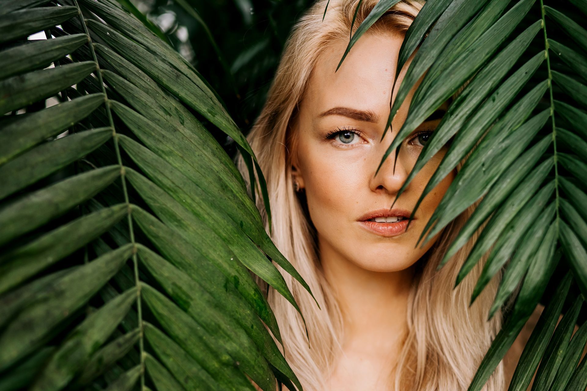 Blonde woman framed by green palm leaves, partially covering her face, in a lush tropical setting