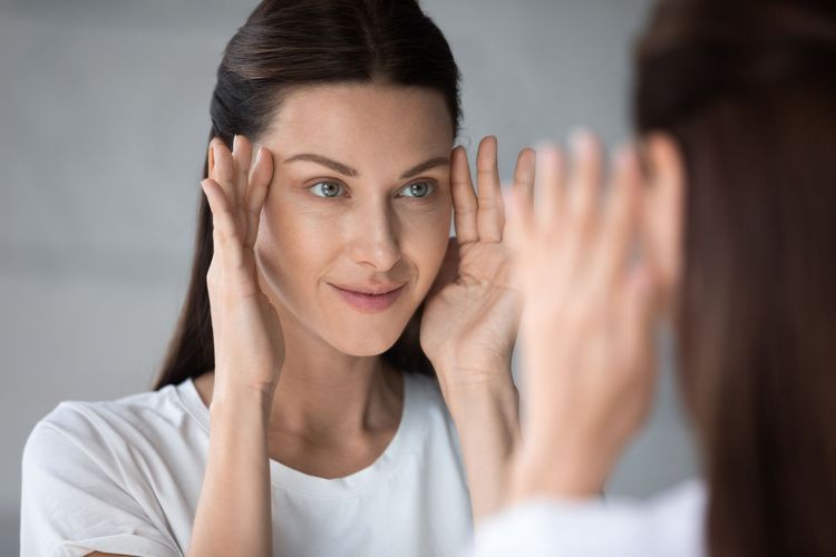 Woman looking at her reflection in a mirror, touching her face in a bright room