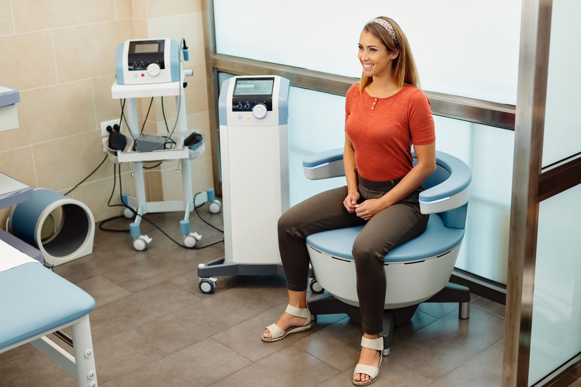 Woman seated on a blue medical device in a clinic room, with equipment nearby.