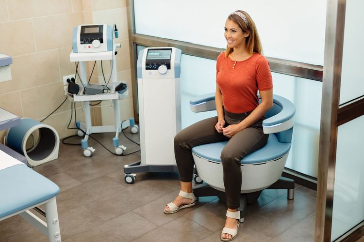 Woman seated on a blue exam chair in a medical office, smiling beside medical equipment.